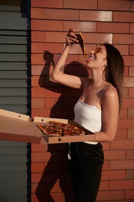 Mulher comendo uma fatia de pizza na rua enquanto segura a caixa da pizza com a outra mão